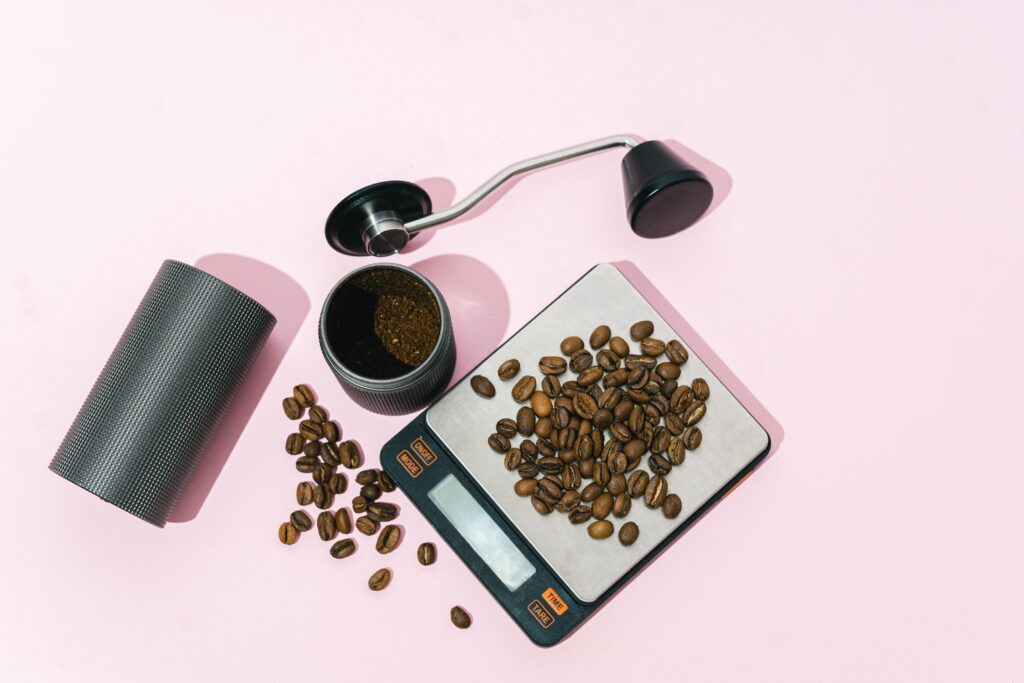 Top view of coffee beans on a digital scale with a grinder on a pink background.